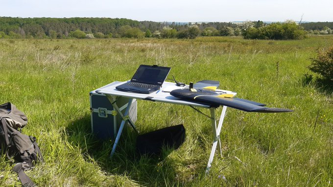 Picture: The photo shows an uncut meadow in the sunshine. In the grass is a folding table on which a laptop and a fixed-wing drone are lying. On the left behind the table is a transport box. On the left at the edge of the picture is a large backpack. Further back in the meadow are groups of bushes and trees and behind them a deciduous forest.