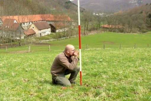 Abbildung: Das Foto zeigt einen auf einer Wiese knieenden Mann neben einem rot und weiß gestreiften sogenannten Fluchtstab, der zur Einmessung kleiner Teilflächen der Experimentier-Plots benötigt wird. Im Hintergrund sind ein Gehöft und Berge zu sehen.