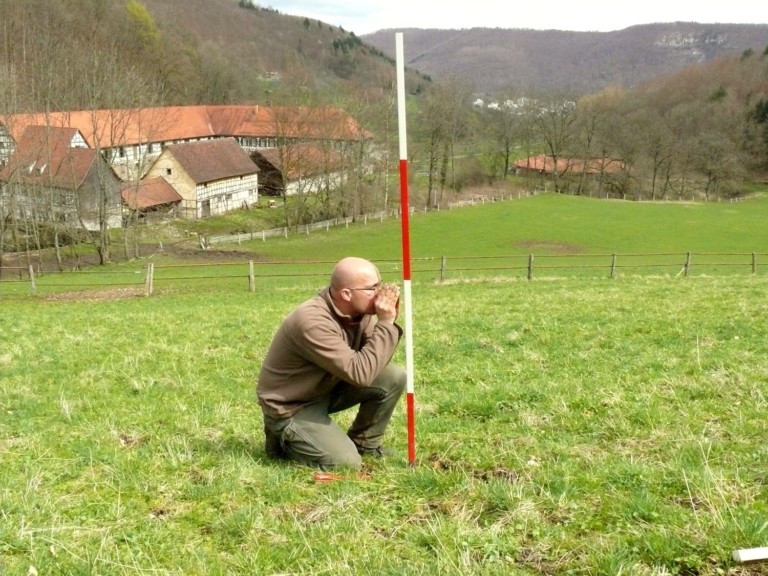 Abbildung: Das Foto zeigt einen auf einer Wiese knieenden Mann neben einem rot und weiß gestreiften sogenannten Fluchtstab, der zur Einmessung kleiner Teilflächen der Experimentier-Plots benötigt wird. Im Hintergrund sind ein Gehöft und Berge zu sehen.