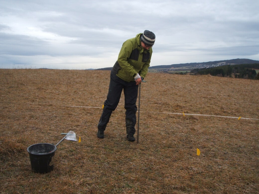 Picture: The photograph shows a scientist in a meadow in winter, using both hands to push an earth boring stick into the ground to take soil samples. Next to the man on the ground are a black plastic bucket and another earth boring stick and a plastic bag.