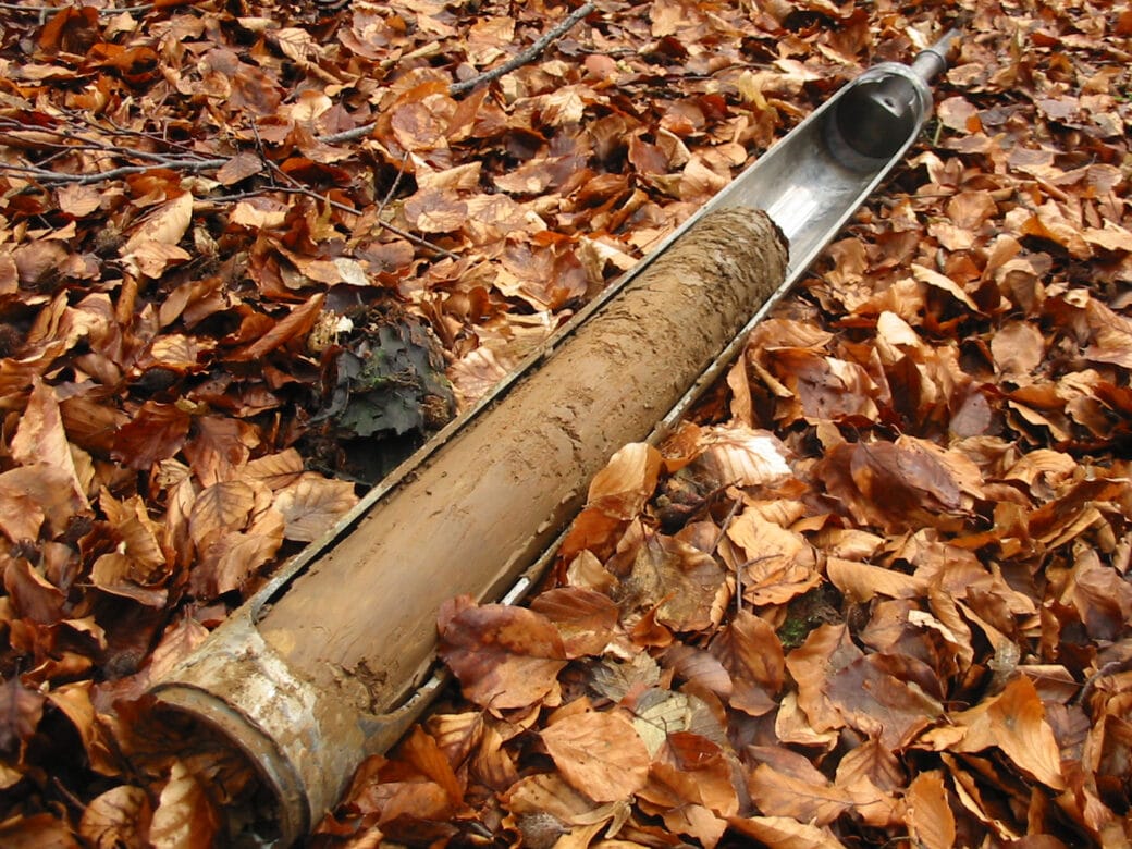 Figure: The photo shows an earth drill stick lying on a forest floor covered with foliage. The container of the drill stick contains a long soil sample.