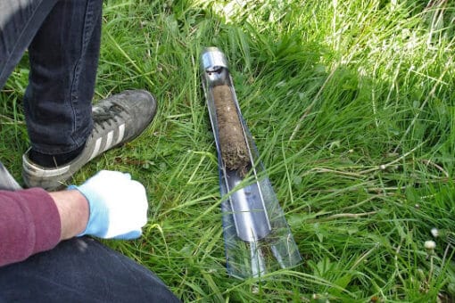 Abbildung: Das Foto zeigt den Behälter eines Erdbohrstocks, der im hohen Gras einer Wiese liegt. Der Behälter enthält eine Bodenprobe.