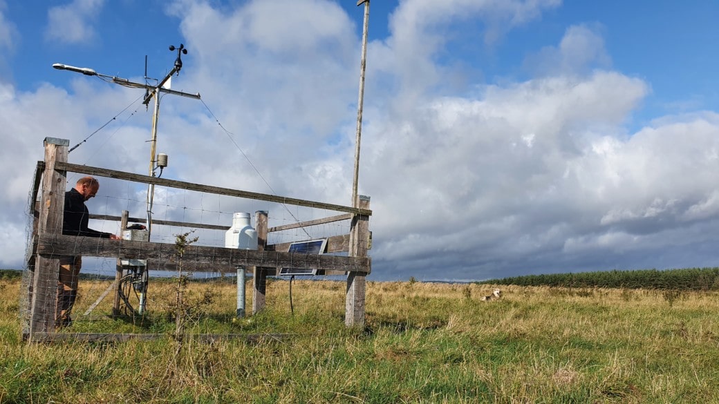 Abbildung: Das Foto zeigt einen Messtechniker bei der Bedienung einer umzäunten Klimamess-Station auf freiem Feld unter blauem Himmel mit Wolken.