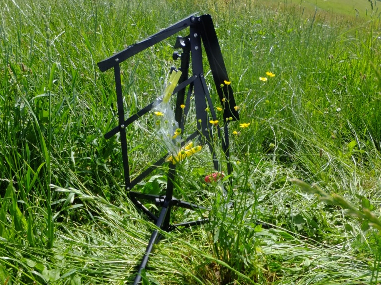 Picture: The photo shows on an uncut meadow in the sunshine an erected black music stand, which has been misappropriated for collecting the scents of flowers and leaves. The tray for the sheet music hangs downward. Attached to the struts of the tray with yellow clothespins are roasting tubes made of transparent plastic film. In the tubes are short pieces of silicone tubing, to the surfaces of which the plant scent molecules adhere. In the upper left of the photo, the corner of a fenced climate measurement station juts into the picture. In the background is a meadow on a slope, on which a row of deciduous trees can be seen above.