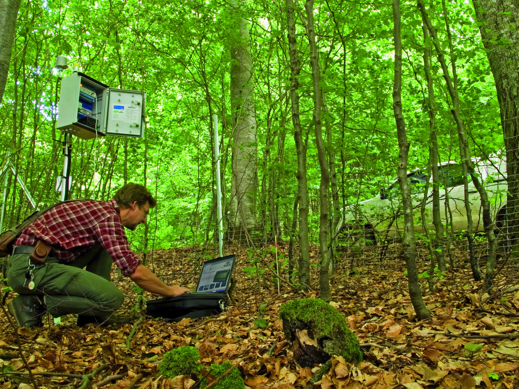 Abbildung: Das Foto zeigt in einem schattigen Wald einen knieenden Mechatroniker an einem Läpptopp beim Auslesen der Daten einer Mess-Station.