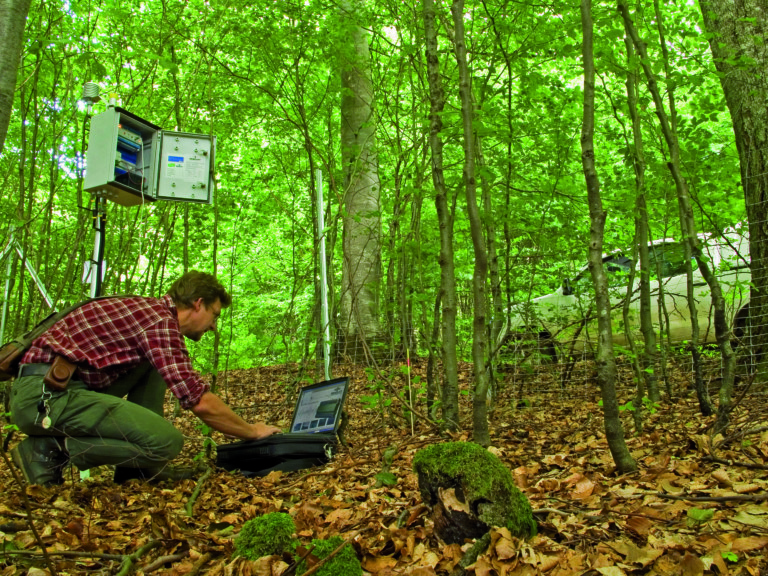 Abbildung: Das Foto zeigt in einem schattigen Wald einen knieenden Mechatroniker an einem Läpptopp beim Auslesen der Daten einer Mess-Station.