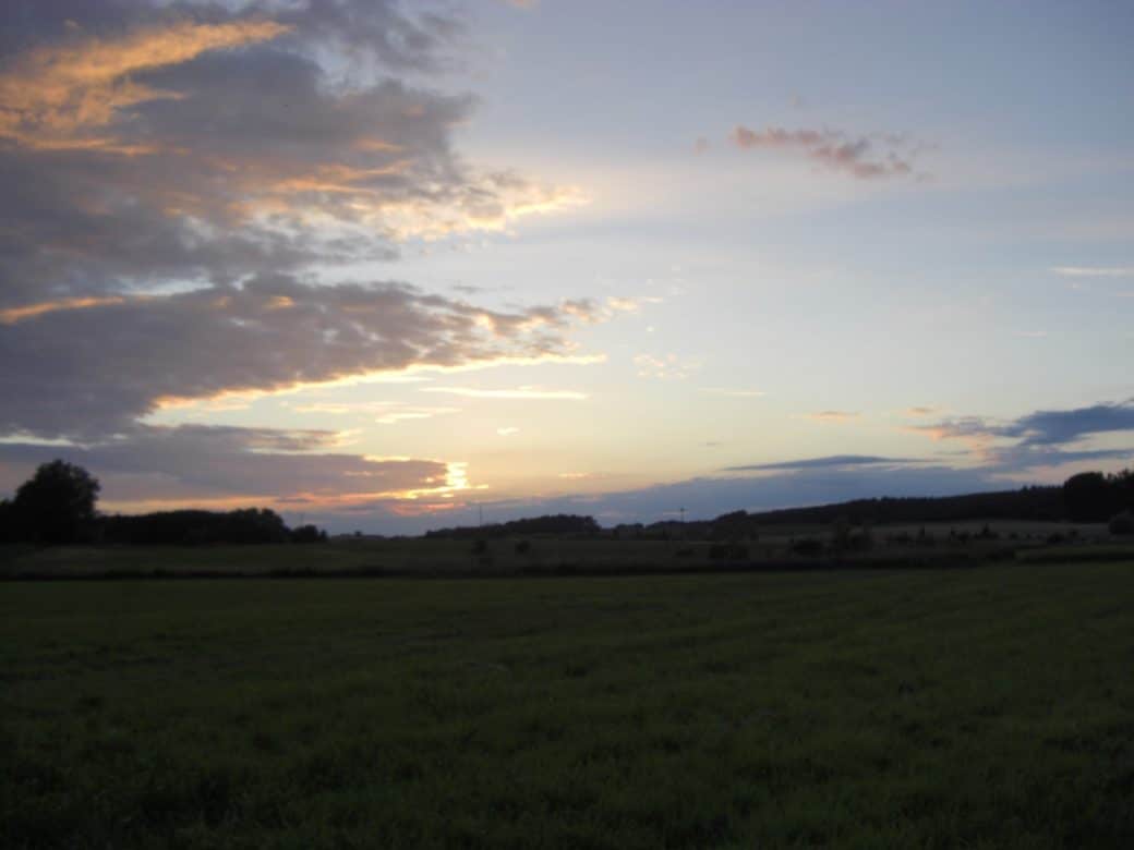 Picture: The photo shows a dark meadow at dawn or dusk, with forested hills on the horizon. The blue sky is partially covered by stratus clouds with an orange-red glow at the edges. The sun is above the horizon hidden behind clouds.