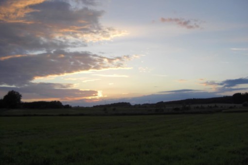 Picture: The photo shows a dark meadow at dawn or dusk, with forested hills on the horizon. The blue sky is partially covered by stratus clouds with an orange-red glow at the edges. The sun is above the horizon hidden behind clouds.