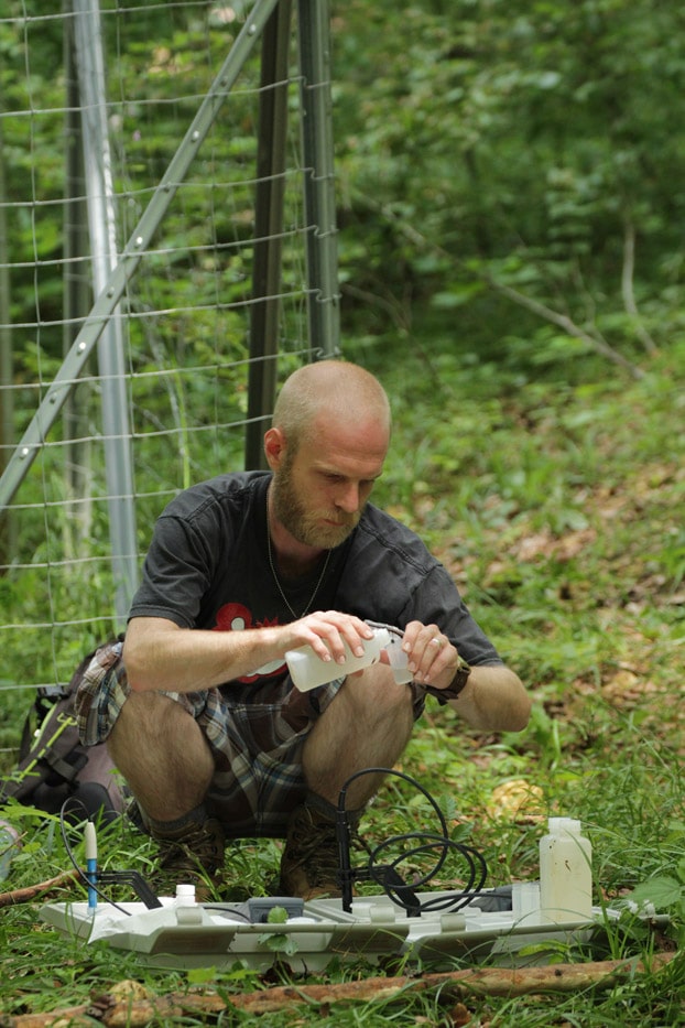 Picture: The photo shows the PhD student Martin Schwarz in a summer forest, squatting on the ground in front of a fenced measuring station and filling a partial sample of precipitation water from a plastic bottle into a small vessel for measuring the electrical conductivity and the P H value. Two plastic trays containing plastic bottles and the measuring device are lying on the ground.