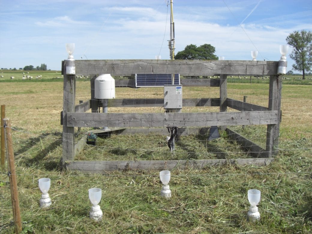 Picture: The photo shows a fenced climate measuring station on a summer meadow. In front of the station, four white plastic bottles are stuck in the ground for about two thirds of their length. Above the screw caps of the bottles, semi-transparent cup-shaped containers open at the top are attached to collect precipitation