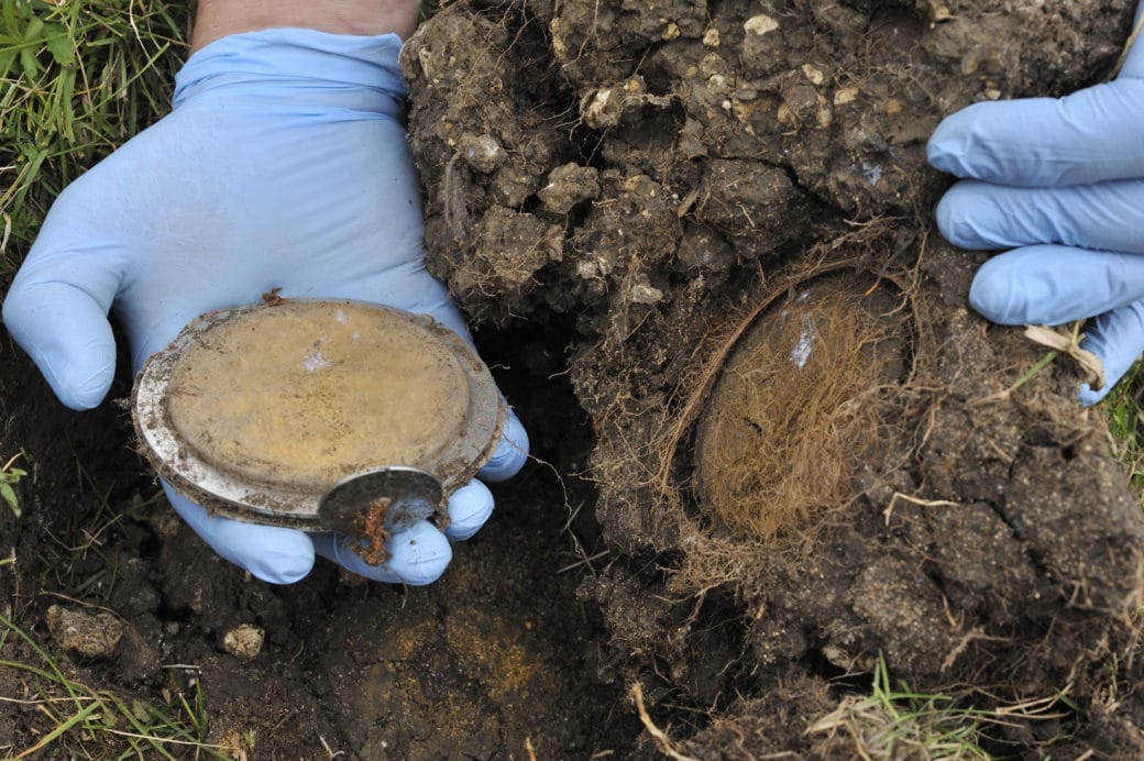 Abbildung: Das Foto zeigt über dem Boden einer Wiese links im Bild eine Handfläche, auf der ein Mineral-Container liegt. Die Hand steckt in einem blauen Latex-Handschuh. Rechts im Bild sieht man eine ebenfalls behandschuhte Hand, die ein großes Stück Erdkrume festhält, das auf die Seite gedreht wurde. Unten an der Krume befindet sich eine kreisförmige, vertiefte Stelle, an der sich der Mineral-Container befand. Innerhalb des Kreises ist ein Geflecht vieler feiner Wurzeln sichtbar.