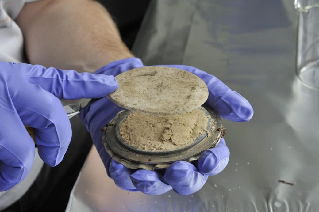 Picture: The photo shows the construction of a mineral container for spreading or installing mineral mixtures in the soils of forests and grasslands. In his left hand, a scientist holds the round, flat metal container for the mixture. The container has a diameter of about ten centimetres and is about one centimetre high. It consists of two discs screwed together with a layer of permeable material between them. Approximately one centimetre from the edge of the upper disc is a circumferential elevation of a few millimetres in height and thickness. The mineral mixture is placed inside the elevation. The mixture is fixed by a permeable textile cap that is pulled over the elevation. In the picture, the scientist's right hand can be seen holding the pulled-off cap between his index finger and a knife blade. Both hands are in blue latex gloves.