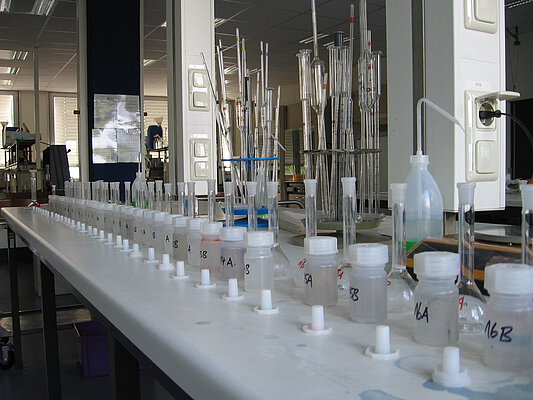 Picture: The photo shows a long table in a large laboratory room, on which containers for a series of measurements for the photometric determination of phosphorus concentration are arranged in two rows. In the first row are wide-necked plastic bottles, in the second row are glass volumetric flasks. Behind the flasks are two stands with long, thin, tubular glass containers.