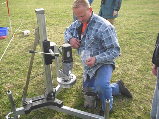Picture: The photo shows a metal device for taking undisturbed soil samples on a mown meadow. The lower part of the device consists of three struts which, viewed from above, form the shape of a ypsilon. Where the struts meet in the middle, a tripod column projects upwards. The motor-driven unit is attached to the stand in a height-adjustable manner. The cutting sleeve is attached to the unit for pre-cutting the sample and the piercing cylinder is attached to it in a fixed position for picking up the sample. Next to the device kneels an employee of the company renting the drill. The stainless steel cutting sleeve is on the floor in front of the man. In his left hand, the man holds a clear transparent piercing cylinder made of acrylic glass.