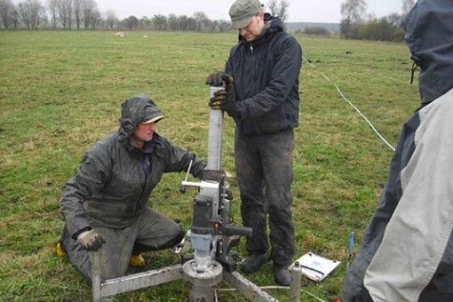 Picture: The photograph shows two men on a large mown meadow operating a device for taking undisturbed soil samples in rainy weather. One of the men is kneeling next to the device, the other is standing and pressing on the tripod column from above. To the right at the edge of the picture another person can be seen watching from behind. A row of unfoliaged trees can be seen on the horizon.