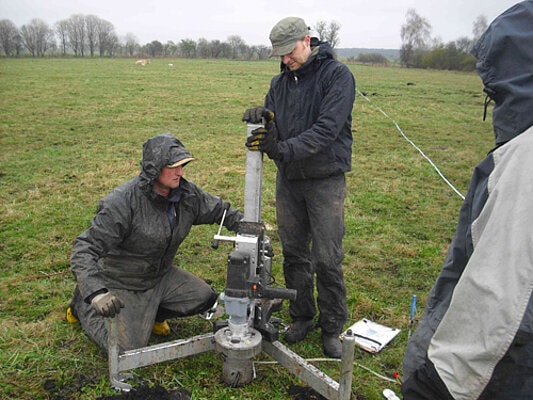 Picture: The photograph shows two men on a large mown meadow operating a device for taking undisturbed soil samples in rainy weather. One of the men is kneeling next to the device, the other is standing and pressing on the tripod column from above. To the right at the edge of the picture another person can be seen watching from behind. A row of unfoliaged trees can be seen on the horizon.