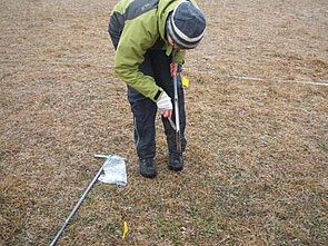 Picture: The photo shows a scientist in a meadow in winter, using a knife to loosen the soil from the cavity of an earth drill stick for taking soil samples. Next to the man, another earth drill stick and a plastic bag lie on the ground.