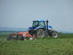 Abbildung: Das Foto zeigt an einem sonnigen Tag einen großen blauen Traktor mit angehängtem Mähwerk beim Mähen einer Wiese.