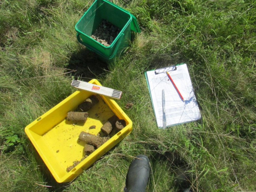 Picture: The photo shows a meadow on which three objects are standing and lying: first, a bucket with small stones in it; second, a yellow bowl containing soil samples with a folding ruler lying on its corner; and third, a clipboard with a form on which a pencil and a knife lie.