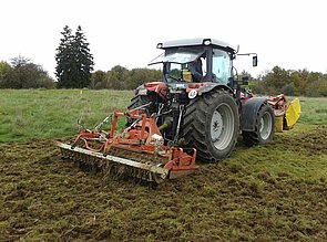 Abbildung: Das Foto zeigt einen Traktor mit angehängtem Gerät beim Aufreißen der Wiesen-Oberfläche zur Verursachung einer Störung.