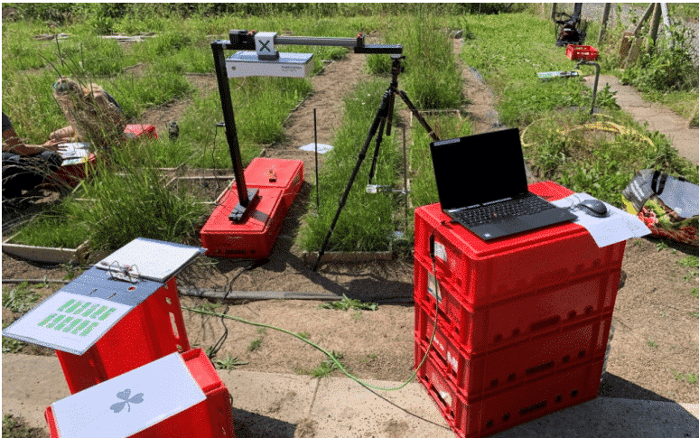 Picture: The photo shows the experimental garden where a multi-spectral 3 D scanner of the brand "Plant Eye" is set up. The scanner stands on an inverted flat red plastic transport box on one of the paths between the sub-plots. The shape of the scanner is similar to that of a crane or gallows: a horizontal boom is attached to a vertical tube, to which the scanning unit is attached and can be positioned differently. The scanning unit is about the size of a shoebox. The scanner's boom extends to the right and rests on top of the plate of a three-legged photo tripod for support. On the next path to the left of the scanner, two young female scientists sit on upturned red transport boxes and study documents. In the front right of the picture, four more upturned red transport crates stand on top of each other and serve as a platform. On the top crate is a laptop with a computer mouse next to it. A cable from the laptop leads to the scanner. In the front left of the picture, two of the red crates are standing on end and serve as storage surfaces for an opened folder and a transparent envelope with documents in it.
