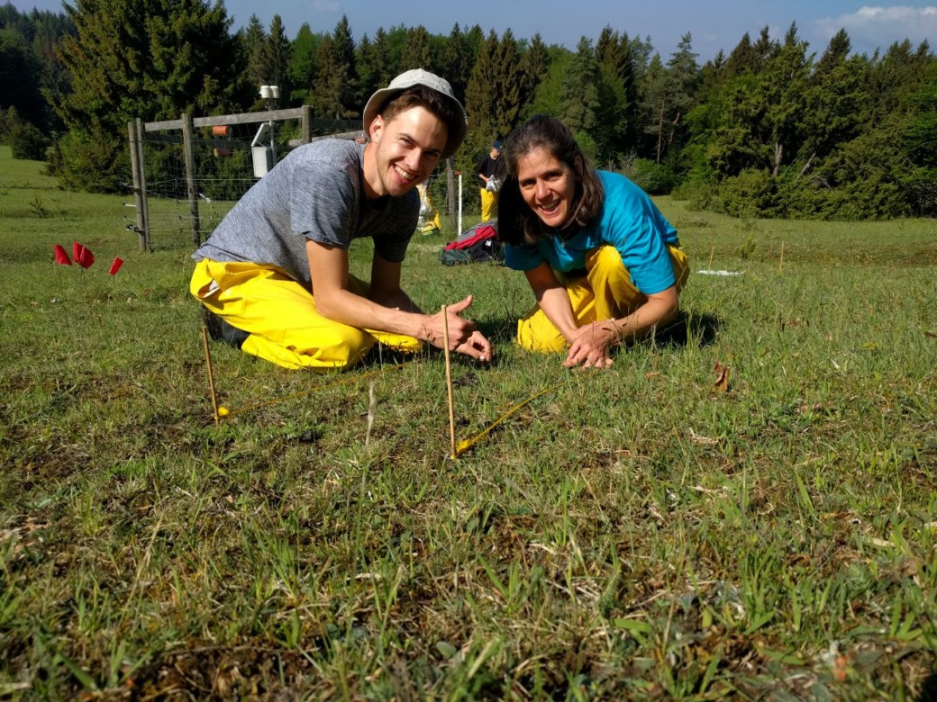 Picture: The photo shows a meadow with a female scientist and a male scientist kneeling on the ground and laying down markers. Both persons are smiling into the camera, in the background a climate measuring station and a forest can be seen.