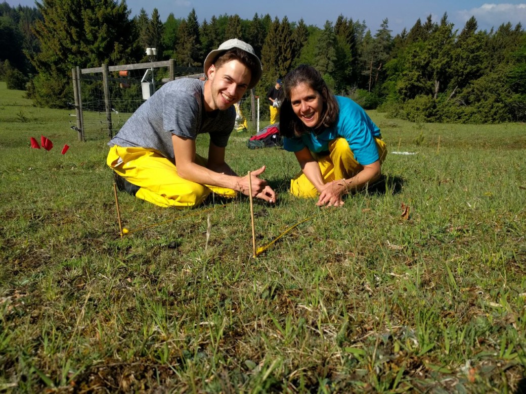 Abbildung: Das Foto zeigt eine Wiese mit
mit einer Wissenschaftlerin und einem Wissenschaftler, die auf dem Boden knien und mit dem Anlegen von Markierungen beschäftig sind. Beide Personen lächeln in die Kamera, im Hintergrund sind eine Klimamess-Station und ein Wald zu sehen.