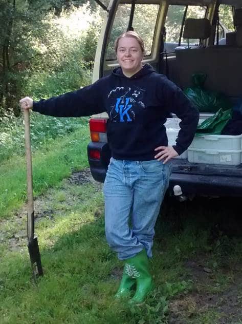 Picture: The photo shows a young female scientist smiling into the camera, standing in an energetic pose in front of the open hatch of a box truck. She is leaning on a spade with her right arm and her left arm is resting on her hip.