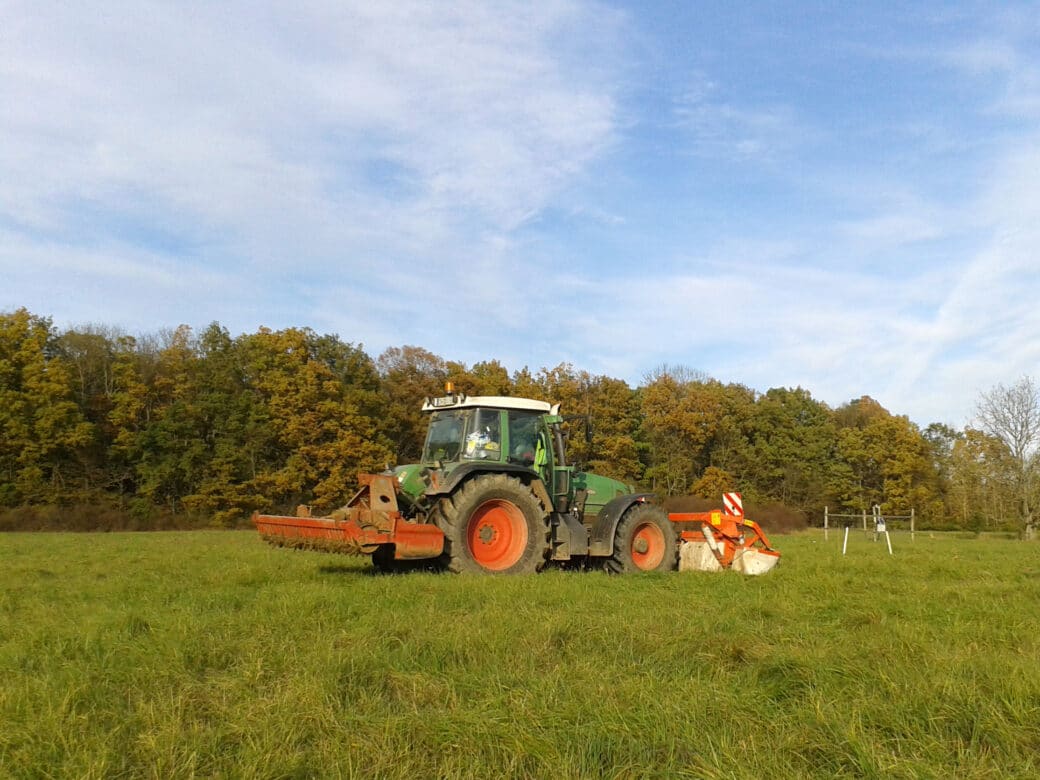 Abbildung: Das Foto zeigt unter blauem Himmel mit Schleierwolken einen Traktor auf einer ungemähten Wiese. Im Hintergrund ist ein Laubwald zu sehen. Die Blätter der Bäume sind herbstlich verfärbt.