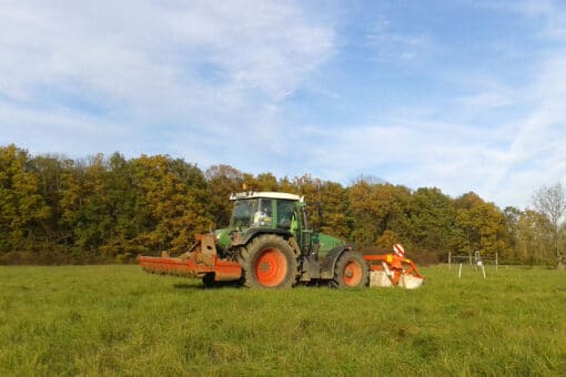 Illustration: The photo shows a tractor on an uncut meadow under a blue sky with veil clouds. In the background a deciduous forest can be seen. The leaves of the trees are autumnally colored.