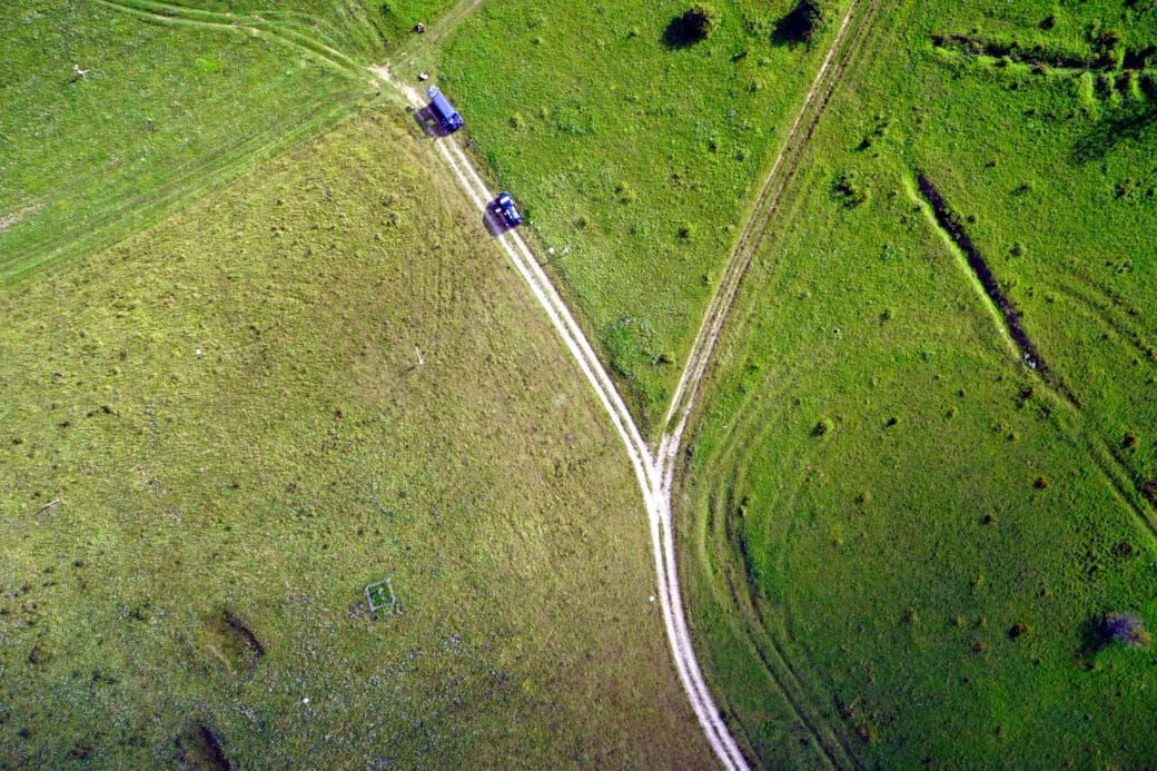 Figure: The drone shot shows a landscape of green meadows with dirt roads in between, photographed from above. Two blue vehicles can be seen on one of the roads