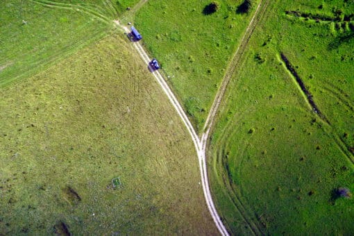 Abbildung: Die Drohen-Aufnahme zeigt von oben fotografiert eine Landschaft von grünen Wiesen mit Feldwegen dazwischen. Auf einem der Wege sind zwei blaue Fahrzeuge zu sehen.