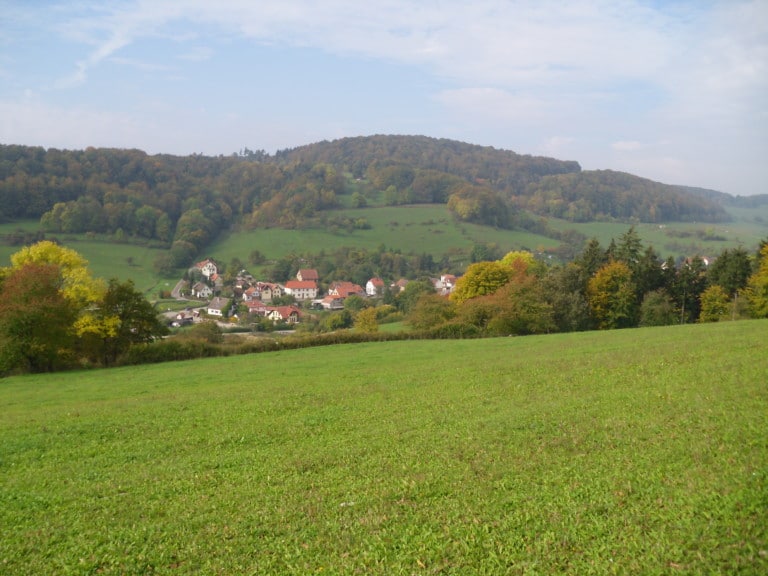 Picture: The photo shows a landscape in autumn with a green meadow in the foreground, a valley with a settlement in the middle and wooded hills in the background.