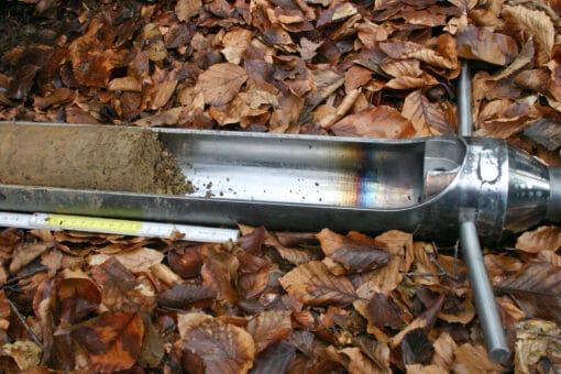 Figure: The photo shows an earth boring stick lying on a forest floor covered with foliage. The container of the boring stick contains a soil sample. A folded-out folding rule lies next to the boring stick.