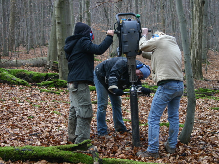 Abbildung: Das Foto zeigt drei Männer im Wald beim Halten und Ausrichten eines Kernbohrgeräts zur Bodenproben-Entnahme.