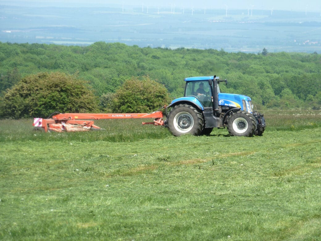 Abbildung: Das Foto zeigt einen Traktor mit angehängtem Gerät beim Mähen einer Wiese. Im Hintergrund ist ein Laubwald zu sehen.