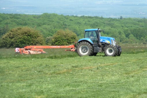 Abbildung: Das Foto zeigt einen Traktor mit angehängtem Gerät beim Mähen einer Wiese. Im Hintergrund ist ein Laubwald zu sehen.