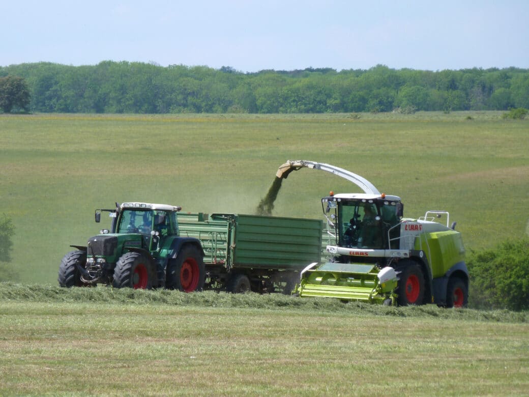 Abbildung: Das Foto zeigt ein landwirtschaftliches Fahrzeug beim Mähen einer Wiese. Über einen Ausleger wird das abgemähte Gras in einen Anhänger befördert, der von einem Traktor gezogen wird, der neben dem anderen Fahrzeug fährt. Im Hintergrund sind eine sehr große Wiese mit einem anschließenden Laubwald zu sehen.