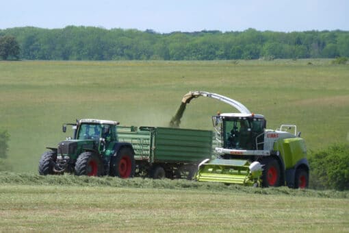 Figure: The photo shows an agricultural vehicle mowing a meadow. Via a boom, the mown grass is transported into a trailer pulled by a tractor driving next to the other vehicle. In the background, a very large meadow with an adjoining deciduous forest can be seen.