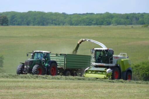 Abbildung: Das Foto zeigt ein landwirtschaftliches Fahrzeug beim Mähen einer Wiese. Über einen Ausleger wird das abgemähte Gras in einen Anhänger befördert, der von einem Traktor gezogen wird, der neben dem anderen Fahrzeug fährt. Im Hintergrund sind eine sehr große Wiese mit einem anschließenden Laubwald zu sehen.