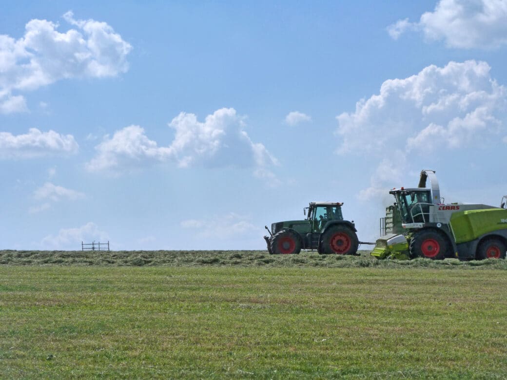 Abbildung: Das Foto zeigt an einem Sommertag eine gemähte Wiese unter blauem Himmel mit einzelnen Wolken. Rechts im Bild sind ein Traktor und ein Mähdrescher zu sehen.