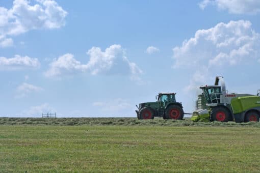 Figure: The photo shows a mowed meadow under a blue sky with single clouds on a summer day. A tractor and a combine harvester can be seen on the right side of the picture.