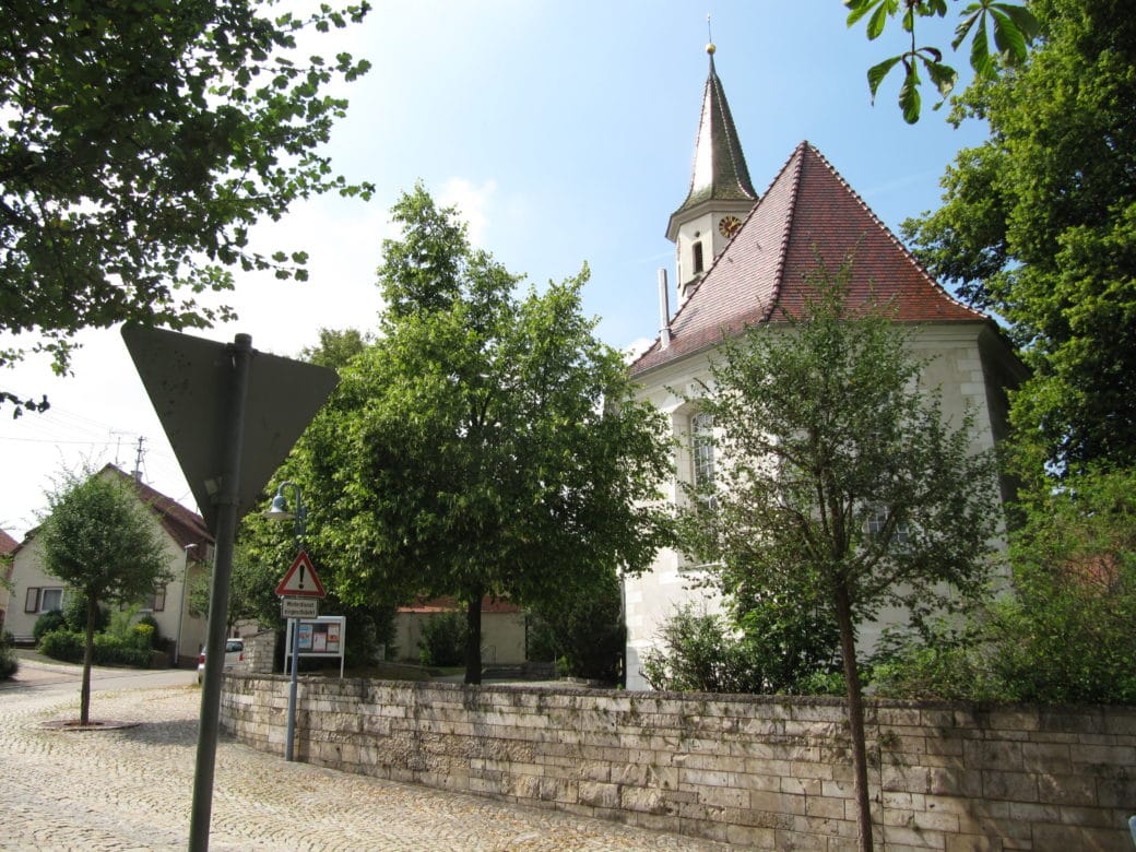 Abbildung: Das Foto zeigt im Ort Kleinengstingen die Kirche und den ummauerten Kirchhof mit Bäumen und Sträuchern darin.