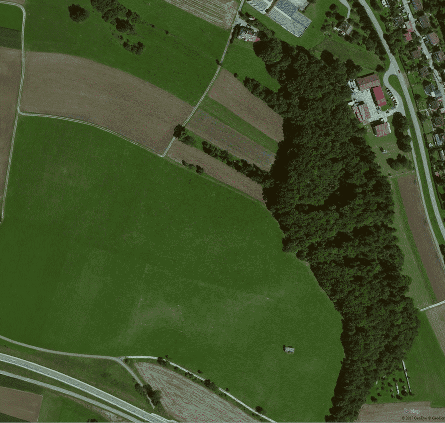 Abbildung: Die Luftaufnahme zeigt grün bewachsene und abgeerntete braune Felder neben einem Waldstück und einer Siedlung.