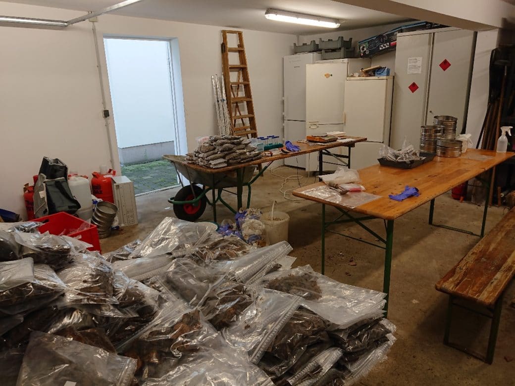 Picture: The photograph shows a deserted room lit by a fluorescent tube on the ceiling. on the left in the background against the wall are canisters, gas bottles, sieves and rucksacks. directly next to it is an open passage to a courtyard. on the right in the background are ladders against the wall. next to them can be seen a metal cupboard and three tall refrigerators or freezers, two of them out of order. in the middle of the room are two long folding tables with wooden table tops. in front of one table is a wheelbarrow, in front of the other a bench. on both tables are various working materials as well as transparent bags with samples in them. in the foreground of the picture can be seen on a table on the left a large heap of bags with samples in them.