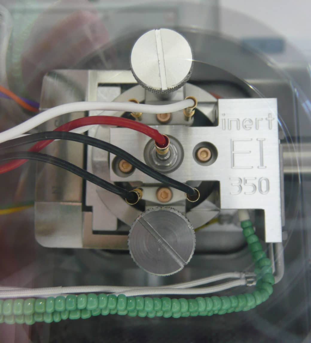 Picture: The photo shows a close-up of a device for electronic ionization photographed from above. You can see a metal housing with a metal holder attached with two large screws, in which a white, a red and two black cables are plugged in. At the bottom of the device there are more cables in different  colors.