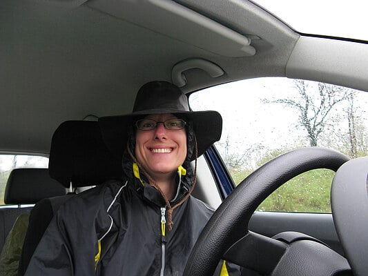 Abbildung: Das Foto zeigt in einem Auto aus Richtung des Lenkrads fotografiert eine in die Kamera lächelnde Wissenschaftlerin mit Funktionskleidung und Hut.