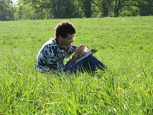 Abbildung: Das Foto zeigt einen jungen Wissenschaftler, der im Sonnenschein auf einer grünen Wiese sitzt. In der rechten Hand hält er eine kleine Pflanze, mit der linken Hand hält er ein aufgeschlagenes Buch fest, das auf seinen Knien liegt.