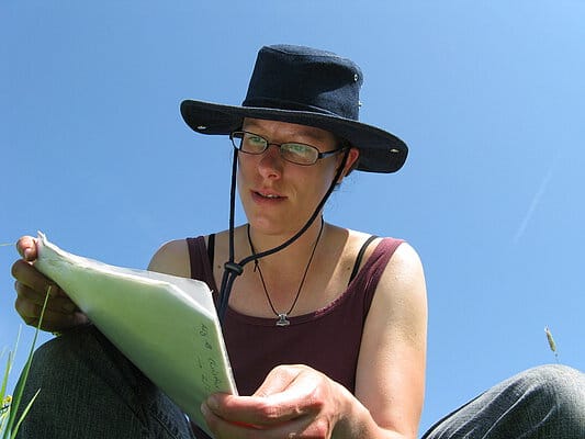 Picture: The photo shows a scientist with a sun protection hat sitting on a meadow in front of a blue sky and reading something from a pile of sheets of paper in the size DIN A 4, which she holds in her hands.