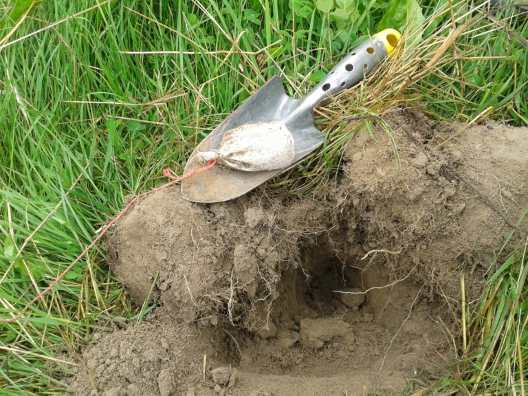 Picture: The photo shows a pile of dug-up earth in a meadow, on which a metal hand shovel is lying, on top of which is a cloth bag filled with exchange resins